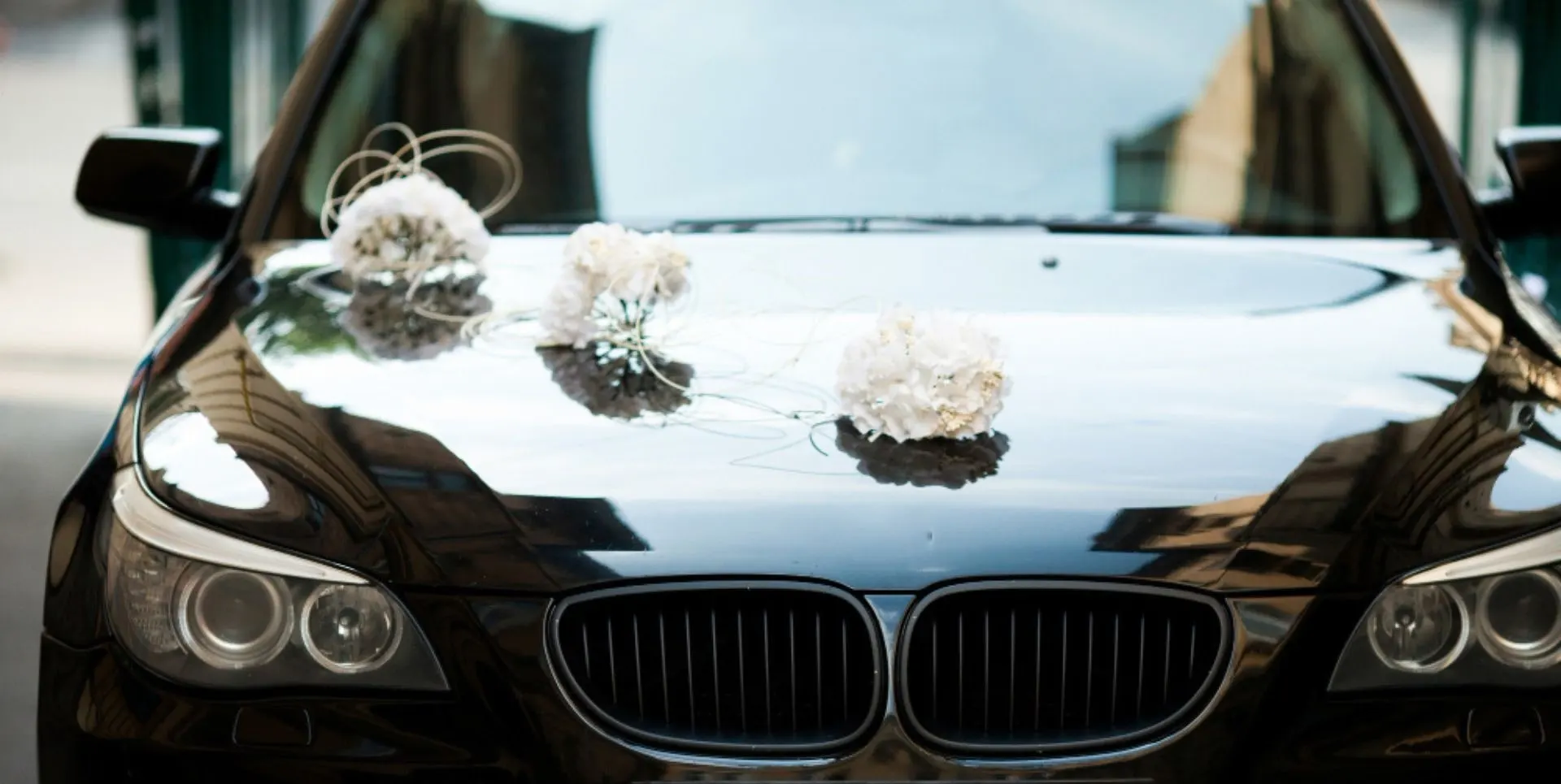 Przód czarnego BMW z charakterystycznym podwójnym grillem i okrągłymi reflektorami. Na masce samochodu znajdują się trzy białe dekoracje ślubne w formie kwiatowych bukietów.