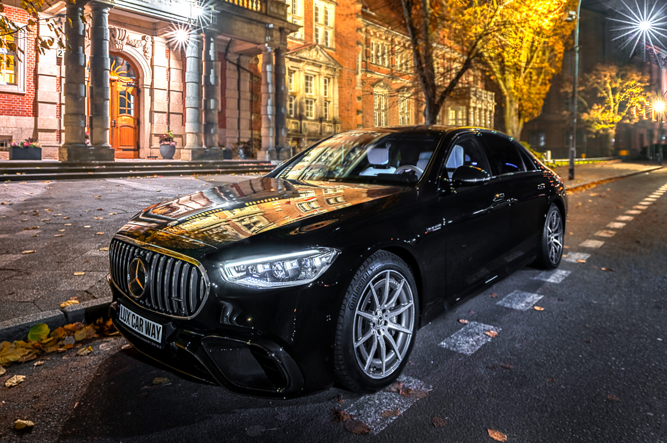 Czarny Mercedes-AMG zaparkowany przed zabytkowym budynkiem z cegły i kolumnami w jesienny wieczór. Oświetlenie uliczne i żółte liście na drzewach tworzą nastrojową atmosferę. Samochód wyróżnia się charakterystycznym grillem AMG i srebrnymi felgami.
