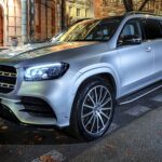 Srebrny Mercedes-Benz GLS zaparkowany przed zabytkowym budynkiem z cegły. SUV wyposażony w wieloramienne felgi aluminiowe i LED-owe reflektory. Jesienne liście widoczne na chodniku.