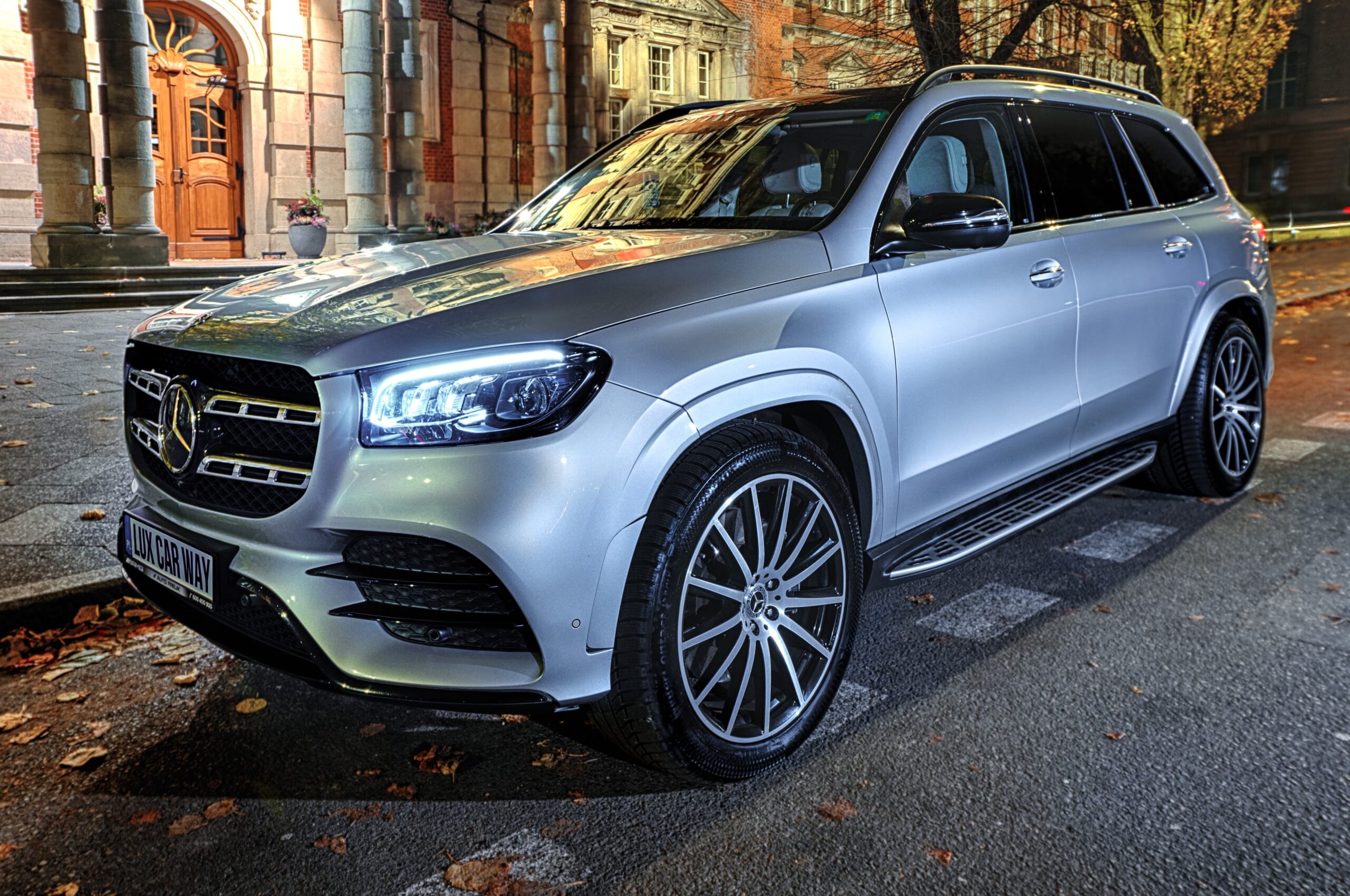 Srebrny Mercedes-Benz GLS zaparkowany przed zabytkowym budynkiem z cegły. SUV wyposażony w wieloramienne felgi aluminiowe i LED-owe reflektory. Jesienne liście widoczne na chodniku.