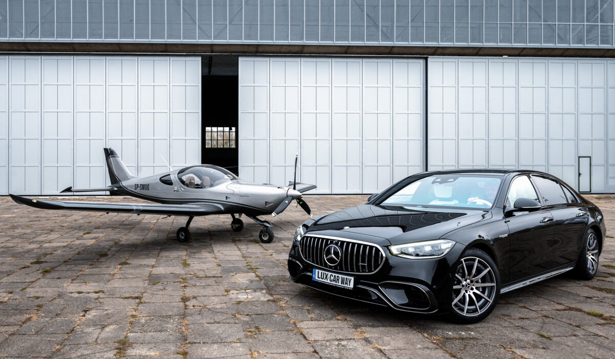 Czarny Mercedes-AMG klasy S zaparkowany przed hangarem lotniczym obok małego srebrnego samolotu sportowego z oznaczeniem SP-SMUG. W tle widoczna biała ściana hangaru z dużymi przesuwnymi drzwiami.