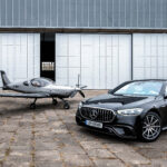 Czarny Mercedes-AMG klasy S zaparkowany przed hangarem lotniczym obok małego srebrnego samolotu sportowego z oznaczeniem SP-SMUG. W tle widoczna biała ściana hangaru z dużymi przesuwnymi drzwiami.