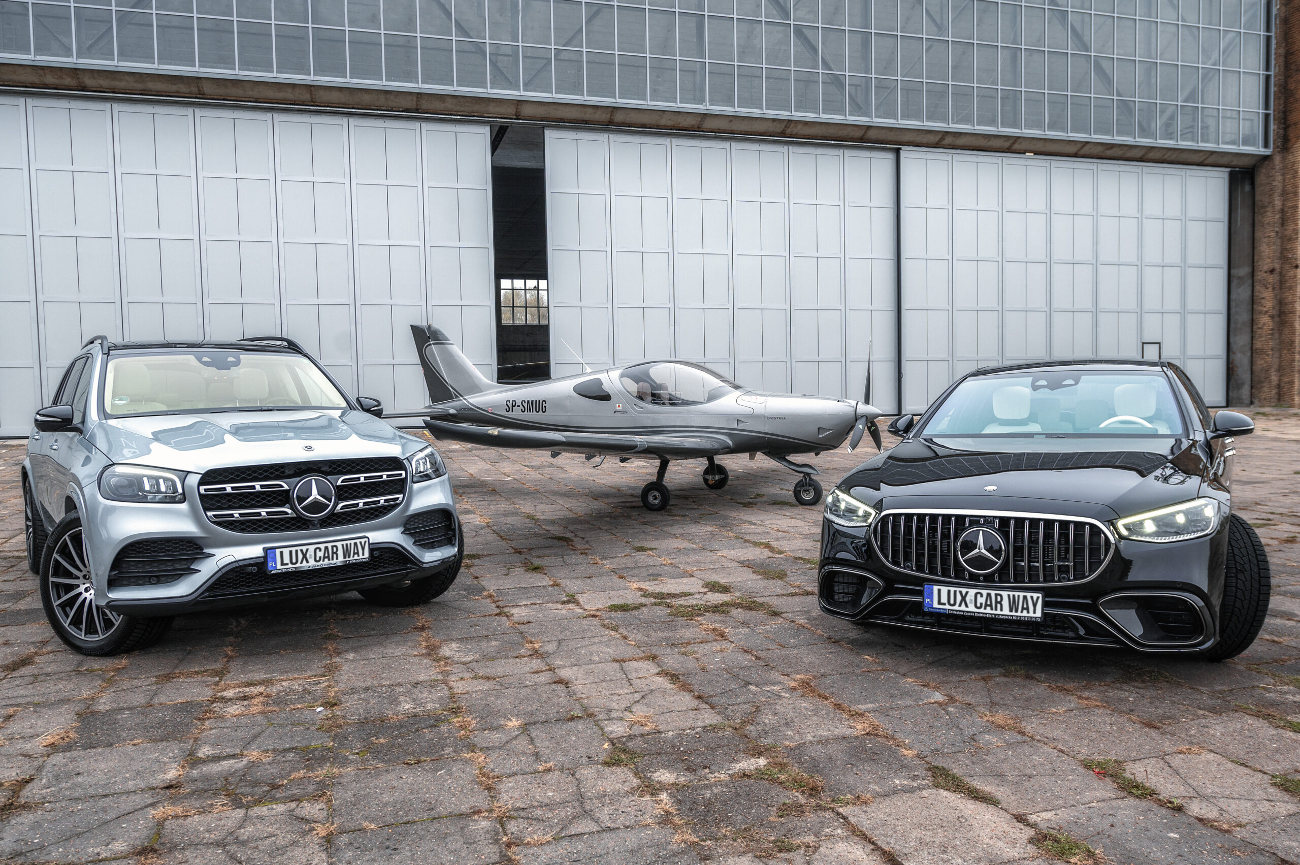 Dwa luksusowe samochody Mercedes-Benz - srebrny GLS i czarny AMG GT - zaparkowane przed hangarem lotniczym. Pomiędzy nimi stoi mały samolot sportowy w kolorze szarym z oznaczeniem SP-SMUG. Wszystkie pojazdy stoją na betonowym placu przed białą ścianą hangaru.