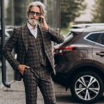 "Elegancko ubrany starszy mężczyzna w kraciaste garniturze i okularach przeciwsłonecznych rozmawia przez telefon przed salonem samochodowym. W tle widoczny jest luksusowy SUV."
