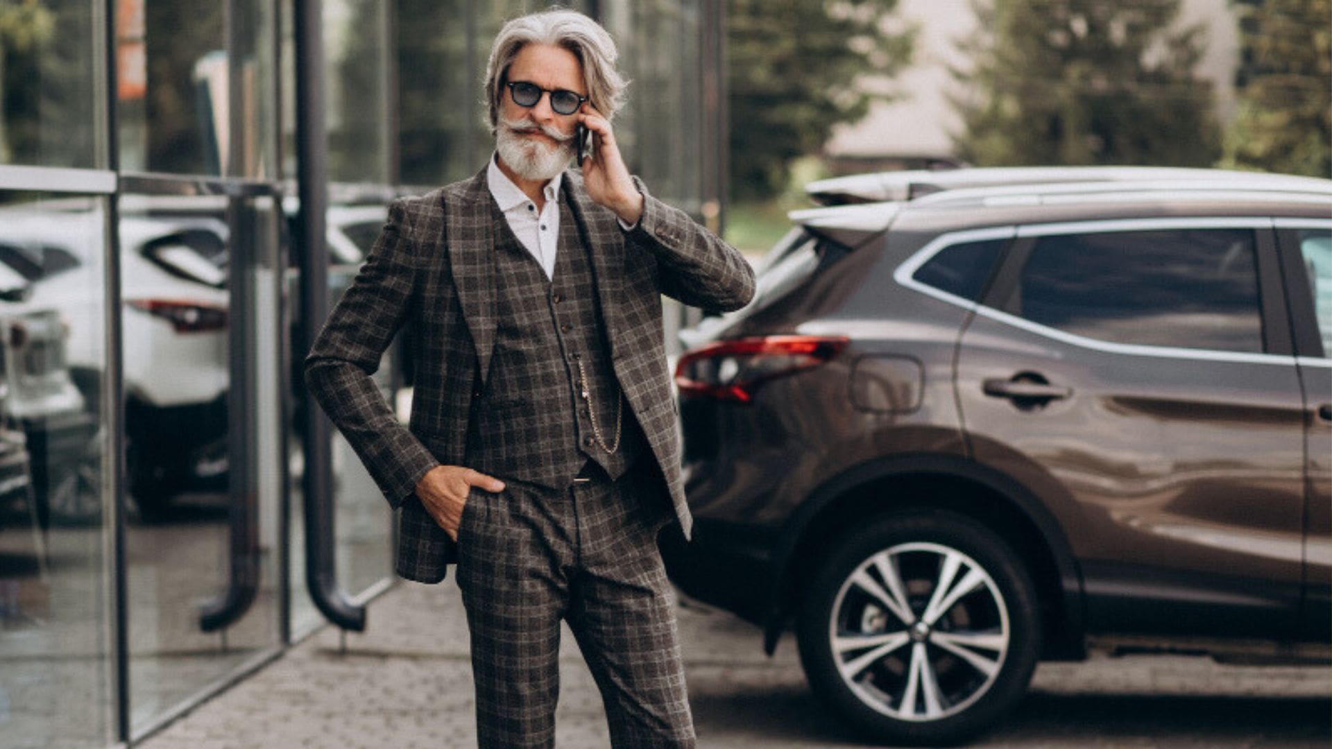 "Elegancko ubrany starszy mężczyzna w kraciaste garniturze i okularach przeciwsłonecznych rozmawia przez telefon przed salonem samochodowym. W tle widoczny jest luksusowy SUV."