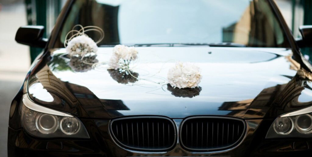 Przód czarnego BMW z charakterystycznym podwójnym grillem i okrągłymi reflektorami. Na masce samochodu znajdują się trzy białe dekoracje ślubne w formie kwiatowych bukietów.