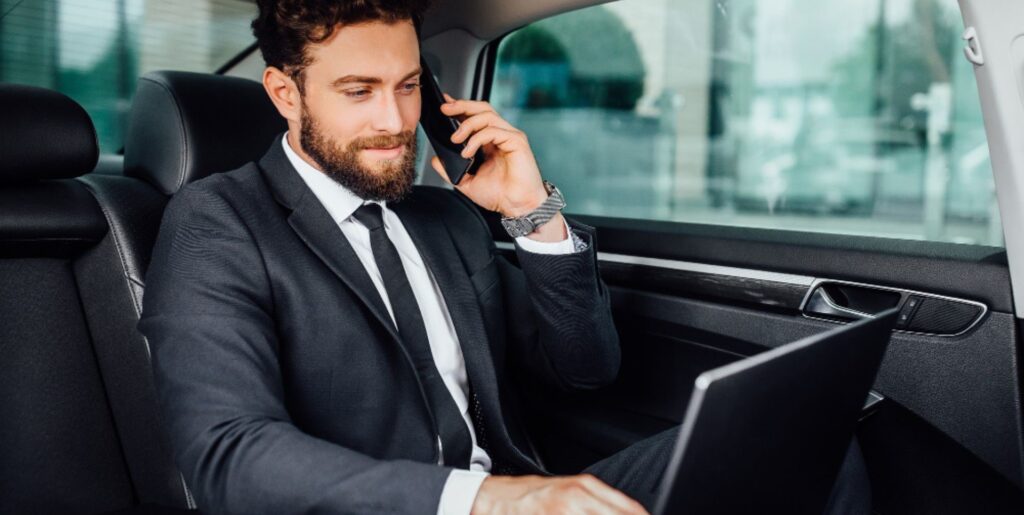 "Biznesmen w czarnym garniturze siedzi na tylnym siedzeniu luksusowego samochodu, rozmawiając przez telefon i pracując na laptopie. Wnętrze pojazdu wykończone jest czarną skórą."