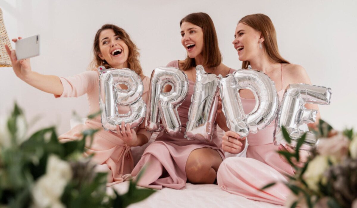 Trzy roześmiane kobiety w różowych sukienkach robią selfie, trzymając srebrne balony foliowe tworzące napis "BRIDE". W tle widoczne białe kwiaty i zielone liście jako dekoracja.