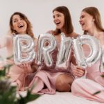 Trzy roześmiane kobiety w różowych sukienkach robią selfie, trzymając srebrne balony foliowe tworzące napis "BRIDE". W tle widoczne białe kwiaty i zielone liście jako dekoracja.