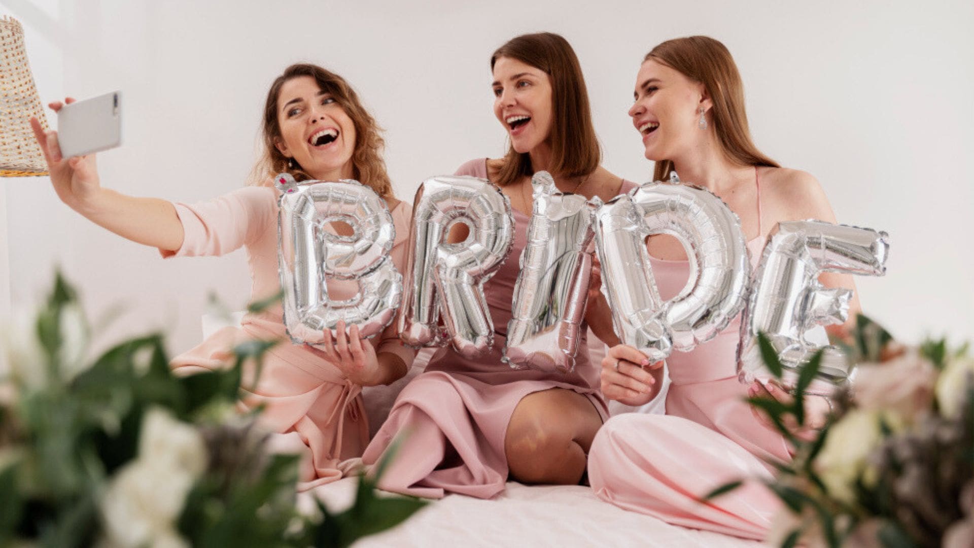 Trzy roześmiane kobiety w różowych sukienkach robią selfie, trzymając srebrne balony foliowe tworzące napis "BRIDE". W tle widoczne białe kwiaty i zielone liście jako dekoracja.